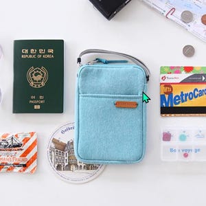 Puede incluir: Una colecci&oacute;n de elementos esenciales de viaje que incluyen un bolso cruzado azul claro, gafas de sol, un pasaporte de la Rep&uacute;blica de Corea, una MetroCard y un peque&ntilde;o organizador de pastillas. Otros art&iacute;culos incluyen un mapa, monedas y un paquete de Fisherman's Friend.