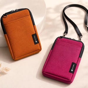 May include: Two rectangular travel pouches, one orange and one magenta, with black trim and zippers. The magenta pouch has a black strap. Each pouch has a small black label with white text. The pouches are on a light-colored surface.