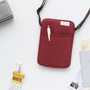 May include: A burgundy felt crossbody bag with a zipper closure and a small pocket on the front. The bag has a black adjustable strap and a white pen is sticking out of the pocket.