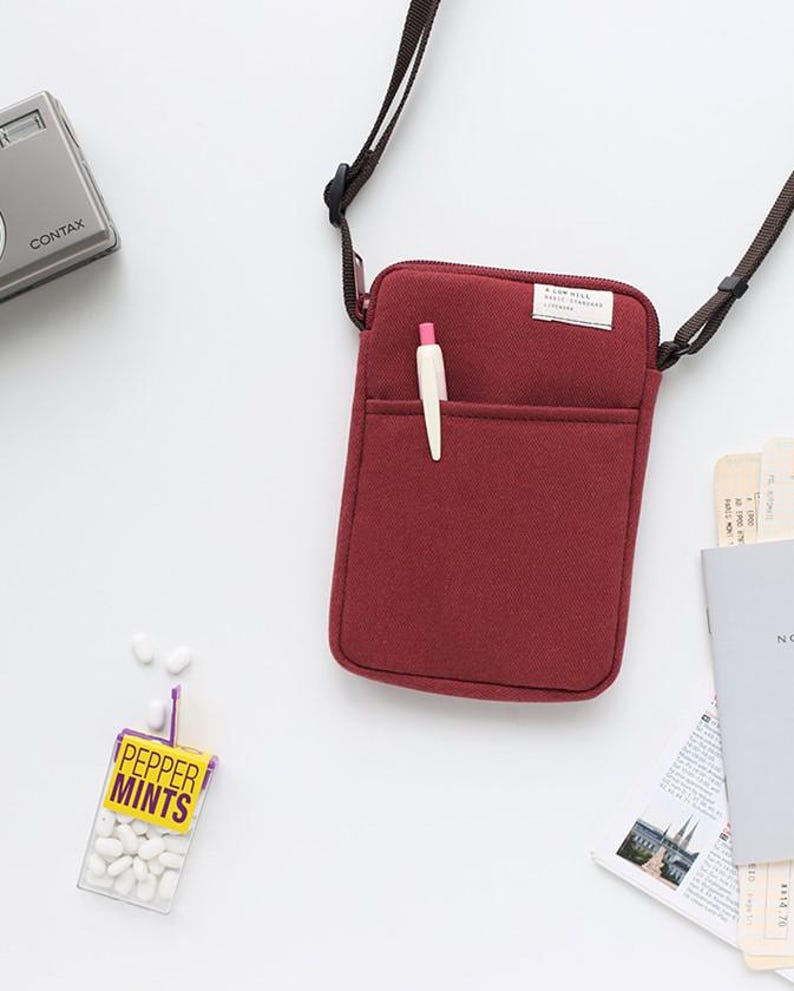 May include: A burgundy felt crossbody bag with a zipper closure and a small pocket on the front. The bag has a black adjustable strap and a white pen is sticking out of the pocket.