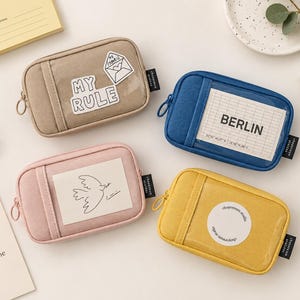 May include: Four rectangular zippered pouches in beige, blue, pink, and yellow. Each pouch features a clear front window displaying unique graphics: a letter with the text MY RULE, a grid with BERLIN, a bird drawing, and a circular logo.