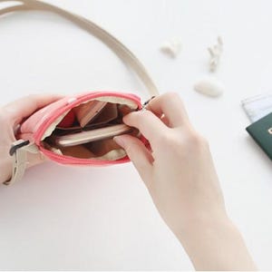 May include: A pink zippered pouch with a beige strap. The pouch is open and a cell phone is inside.