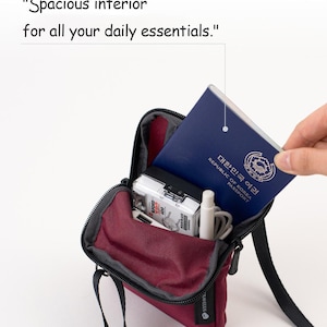 K&ouml;nnte beinhalten: Eine weinrote Umh&auml;ngetasche mit schwarzem Riemen, ge&ouml;ffnet, um einen Reisepass, eine Kamera und andere kleine Gegenst&auml;nde zu zeigen. Die Tasche tr&auml;gt den Text "Spacious interior for all your daily essentials."