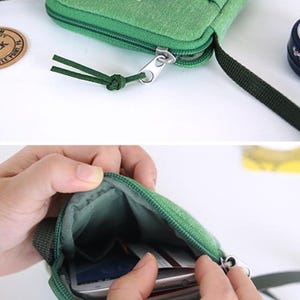 Puede incluir: Bolso bandolera de lona verde con cierre de cremallera y un peque&ntilde;o bolsillo delantero. El bolso tiene una correa verde oscuro y un peque&ntilde;o bol&iacute;grafo y clip decorativos. El interior del bolso es visible, con un smartphone.