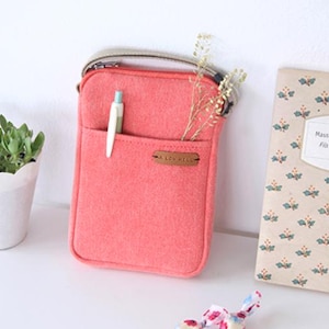 Puede incluir: Un bolso cruzado de fieltro color coral con un bolsillo delantero que contiene un bol&iacute;grafo. El bolso tiene una correa beige y una peque&ntilde;a etiqueta de cuero que dice "A LOV HELLO". Una peque&ntilde;a planta en maceta y un libro con motivos florales tambi&eacute;n est&aacute;n en la imagen.
