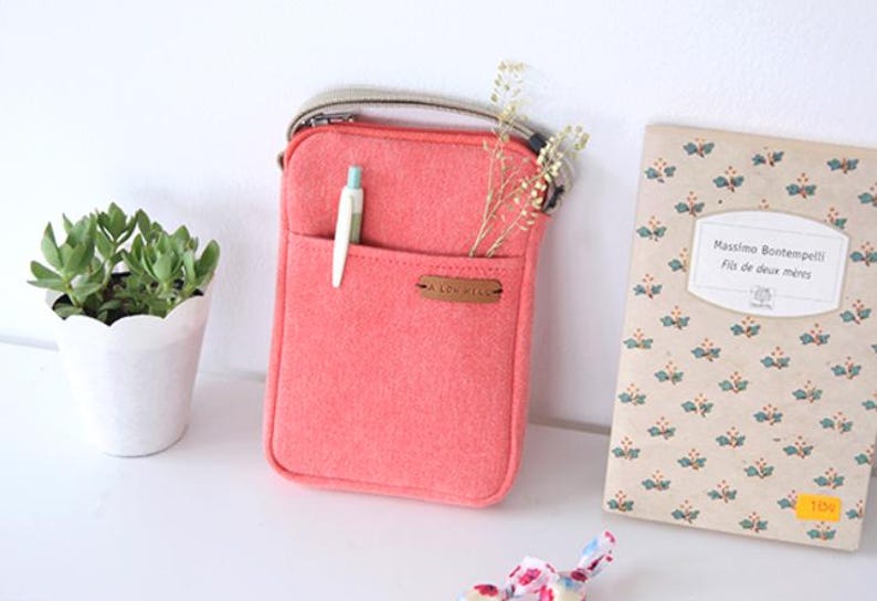 Puede incluir: Un bolso cruzado de fieltro color coral con un bolsillo delantero que contiene un bol&iacute;grafo. El bolso tiene una correa beige y una peque&ntilde;a etiqueta de cuero que dice "A LOV HELLO". Una peque&ntilde;a planta en maceta y un libro con motivos florales tambi&eacute;n est&aacute;n en la imagen.