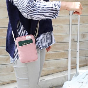 May include: A pink travel wallet with a passport inside, worn on a shoulder strap. The wallet is attached to a white suitcase with a retractable handle.
