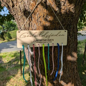 Może przedstawiać: Drewniana tablica z niemieckim tekstem "Geduldsfäden" i "ZUM MITNEHMEN" zawieszona na drzewie. Poniżej tablicy wiszą kolorowe wstążki o różnych długościach. Tablica przymocowana jest do drzewa białym sznurkiem.