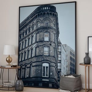 Puede incluir: Interior de dormitorio con una gran impresión enmarcada de un edificio en tonos monocromáticos. A la izquierda, una cama con ropa de cama gris y una almohada blanca. Una pequeña mesita de noche con una lámpara y decoración está al lado de la cama. También se ve una alfombra y una mesa auxiliar con decoración.