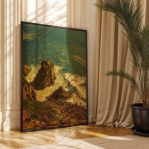 May include: A framed photograph of a rocky cliff overlooking a turquoise ocean with white waves crashing against the shore. The photo is framed in black and is hanging on a wall in a room with a plant and a chair.