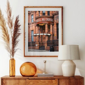 Könnte beinhalten: Ein Holzschrank mit einem gerahmten Bild eines Gebäudes mit der Aufschrift "HOTEL METROPOLE". Der Schrank hat eine Lampe, eine Vase mit getrockneten Pflanzen und eine Glaskugel. Auf dem Boden liegt ein runder Teppich.