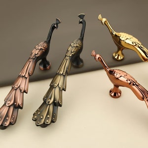 May include: Four peacock-shaped cabinet door pulls in different finishes: gold, copper, bronze, and black. The pulls are detailed with intricate feather patterns.