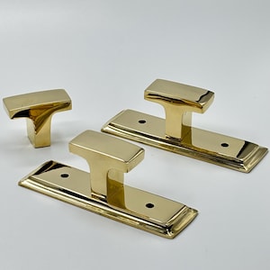 May include: A set of three brass door hardware pieces. Two rectangular backplates with a T-shaped handle and a single T-shaped handle. The hardware has a polished, reflective finish.