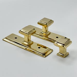 May include: Three brass door pulls with a square knob design. The pulls have a rectangular base with two screw holes. The knobs are a bright, shiny gold colour.