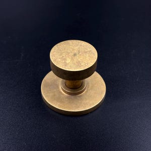 May include: A solid brass door knob with a circular base and a cylindrical top. The knob has a warm, golden hue and a simple, minimalist design. The hardware is likely for a door or cabinet.