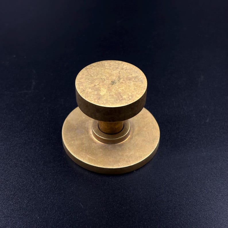 May include: A solid brass door knob with a circular base and a cylindrical top. The knob has a warm, golden hue and a simple, minimalist design. The hardware is likely for a door or cabinet.