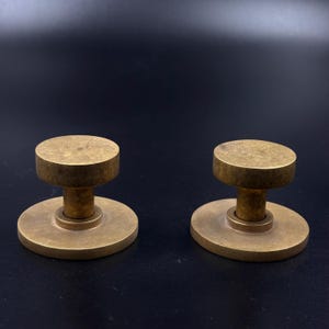 May include: Two brass cabinet knobs with a circular top and base, connected by a short, cylindrical stem. The knobs have a warm, golden hue and are set against a dark background.