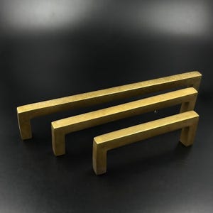 May include: Three brass-toned cabinet pulls in varying sizes. Each pull has a rectangular bar and a squared-off, U-shaped handle. The pulls are arranged in a staggered formation, showcasing their different lengths. The finish has a slightly aged appearance.