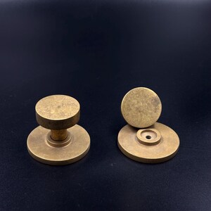 May include: Two antique brass cabinet knobs. One knob is assembled, showing a round base and a cylindrical stem supporting a flat, circular top. The other knob is disassembled, with the top and base separated. The knobs have a warm, aged patina.