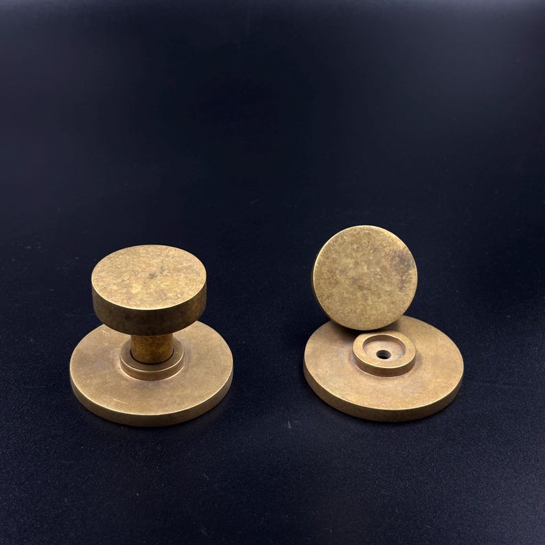 May include: Two antique brass cabinet knobs. One knob is assembled, showing a round base and a cylindrical stem supporting a flat, circular top. The other knob is disassembled, with the top and base separated. The knobs have a warm, aged patina.