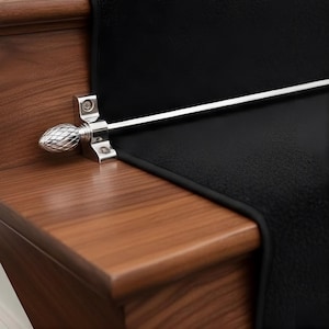 May include: A polished silver stair rod with a decorative finial, installed on a wooden stair step. The rod secures a black stair runner. The finial has a textured, pinecone-like design.