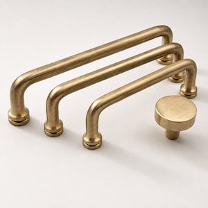 May include: Three elongated, U-shaped metal cabinet pulls with a brushed brass finish and a single matching circular cabinet knob. Each pull features two rounded mounting points at the ends, and the knob has a flat, circular top surface.