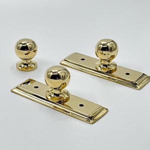 May include: Three gold-coloured cabinet knobs. Two knobs are attached to rectangular plates with rounded edges and two screw holes. The third knob is a standalone sphere-shaped design. All pieces have a shiny, reflective finish.