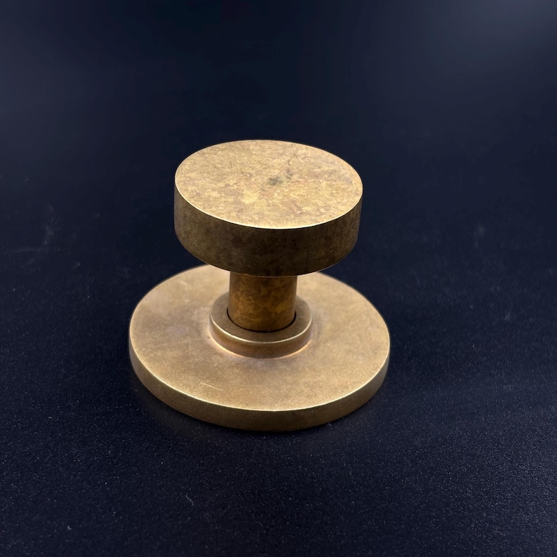 May include: A solid brass door knob with a circular base and a cylindrical top. The knob has a warm, golden hue and a simple, minimalist design. The hardware is likely for a cabinet or door.