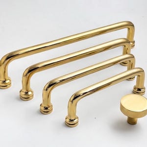May include: A collection of four polished gold-toned metal drawer pulls of varying lengths, each featuring a smooth, rounded handle design with cylindrical mounting bases, accompanied by a single matching circular gold-toned cabinet knob.