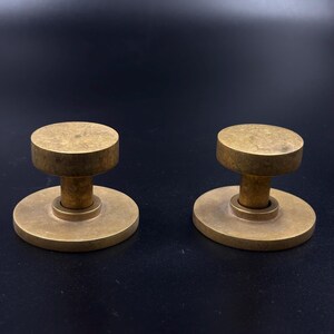 May include: Two brass cabinet knobs with a circular base and a cylindrical top. The knobs have a vintage, antique appearance and are set against a dark background. The knobs are likely for furniture or decorative use.