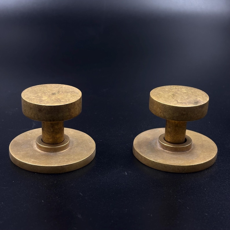 May include: Two brass cabinet knobs with a circular base and a cylindrical top. The knobs have a vintage, antique appearance and are set against a dark background. The knobs are likely for furniture or decorative use.
