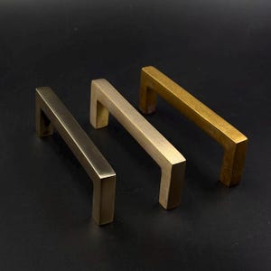 May include: Three brass cabinet pulls in varying shades of gold. The pulls have a rectangular bar shape with right-angle bends at each end, creating a modern, minimalist design. The hardware is set against a dark background.