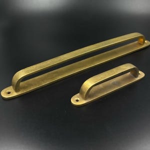 Peut inclure: Deux poignées de porte en laiton. L'une est longue et rectangulaire, l'autre est plus courte. Les deux ont une poignée incurvée et sont montées sur une base rectangulaire avec deux trous de vis. Le métal a un aspect légèrement vieilli.