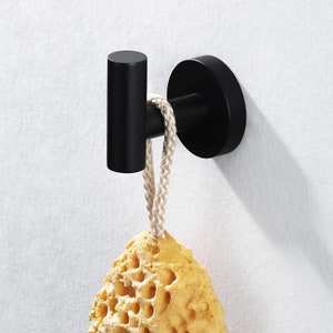 May include: Black metal hook with a round base, mounted on a white wall. A yellow sponge is hanging from the hook with a white rope.