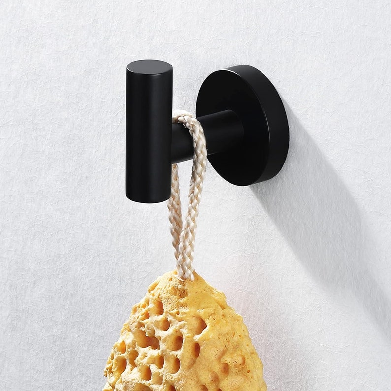 May include: Black metal hook with a round base, mounted on a white wall. A yellow sponge is hanging from the hook with a white rope.