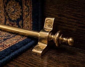 May include: A polished brass stair rod featuring a cylindrical bar and a decorative finial end piece. The metal bracket is secured with two flat-head screws, holding the rod in place against the edge of a blue and gold patterned stair runner.