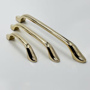 May include: Three gold-coloured cabinet handles of varying sizes are arranged on a white surface. The handles have a curved design with a smooth, polished finish. The largest handle measures approximately 20 cm in length.