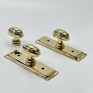 May include: Three polished brass door knobs with oval handles and rectangular backplates. The knobs have a shiny, reflective surface, and the backplates have rounded edges. The hardware is designed for doors.