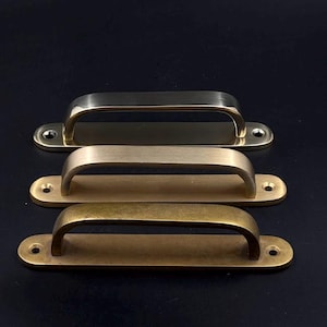May include: Three metal cabinet pulls in varying shades of gold and brass. Each pull has a curved handle and a rectangular base with two screw holes. The top pull is a bright gold, the middle is a lighter brass, and the bottom is a darker, aged brass.