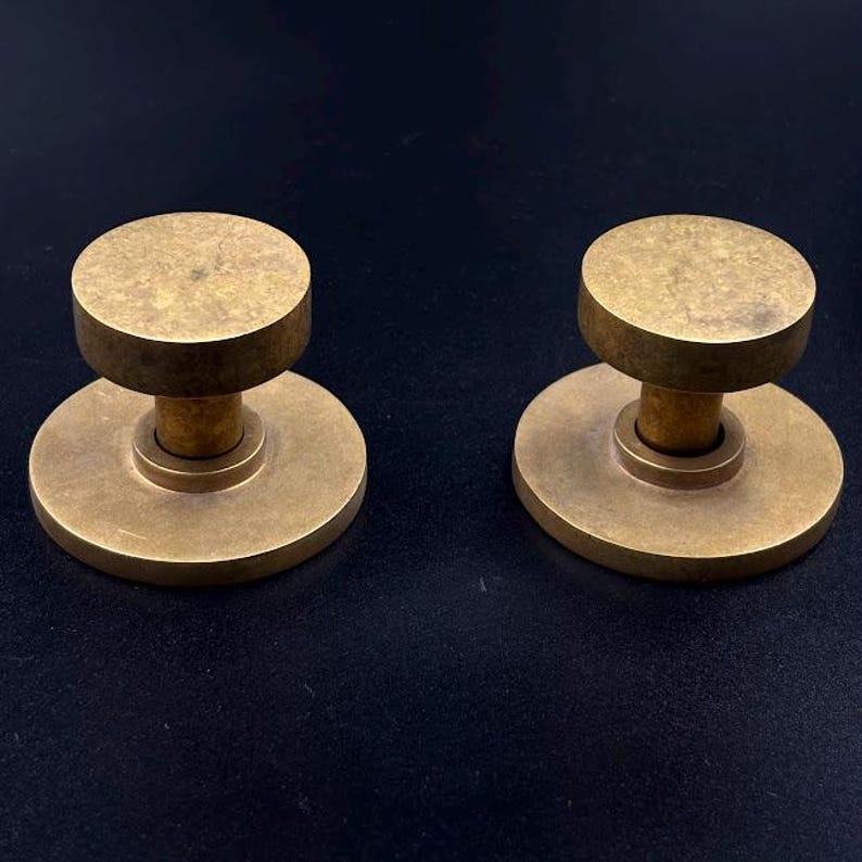 May include: Two brass cabinet knobs with a circular design. Each knob has a flat, round top and a cylindrical stem that connects to a circular base. The knobs are made of a warm, golden-toned metal.