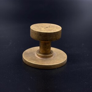 May include: A solid brass door knob with a circular base and a cylindrical top. The knob has a warm, golden hue and a simple, minimalist design. The door knob is likely for a cabinet or drawer.