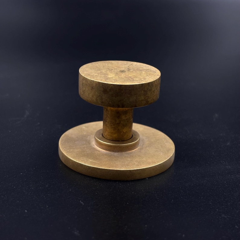 May include: A solid brass door knob with a circular base and a cylindrical top. The knob has a warm, golden hue and a simple, minimalist design. The door knob is likely for a cabinet or drawer.