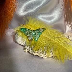 May include: A green butterfly charm with white flowers embedded in the resin. The charm is on a white surface with a yellow feather.