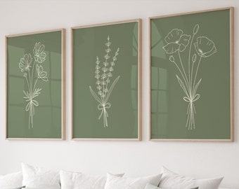 Op de afbeelding: Drie ingelijste prints met een groene achtergrond en witte lijntekeningen van bloemen. De prints tonen een boeket kosmos, een boeket lavendel en een boeket papavers.