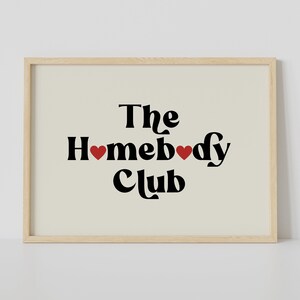 May include: A framed print with the text "The Homebody Club" in black with two red hearts. The background is a light beige.