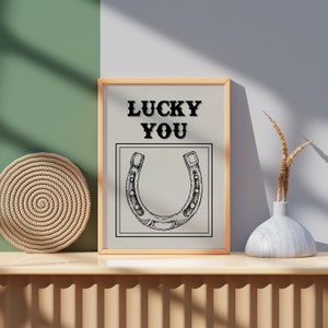 May include: A framed print with a horseshoe illustration and the text "LUCKY YOU" in black lettering on a beige background.
