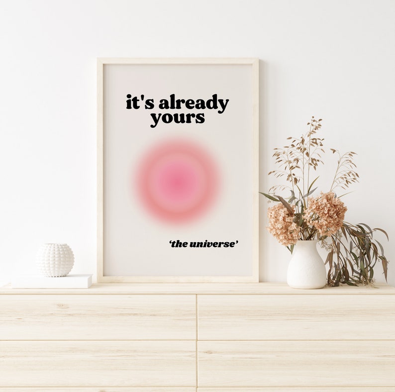 May include: A framed print with a white background and black text that reads "it's already yours" and "'the universe'". A pink and red gradient circle is in the center of the print. The print is hanging on a white wall above a wooden dresser.