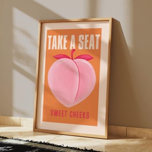 May include: A peach-colored poster with the text "Take a Seat" in white letters and "Sweet Cheeks" in pink letters. A pink peach with a green stem and red leaves is in the center of the poster.