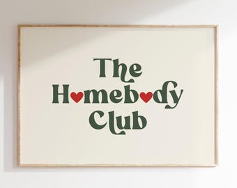 May include: A framed print with the words "The Homebody Club" in dark green, with red heart accents. The print has a cream-colored background and a light brown frame. The text is in a playful, retro font.