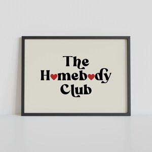 May include: Black and white print with the text "The Homebody Club" in a retro font. Two red hearts are placed between the words "Home" and "body".